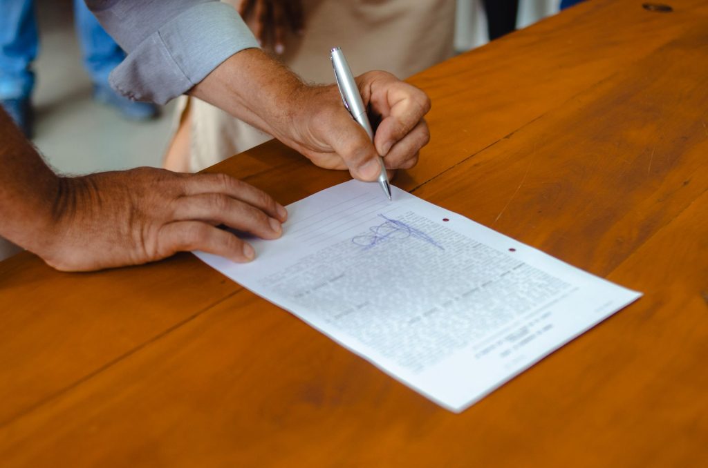A person signing a printed form on a wooden table.
